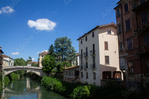 A small bridge and a river next to a street and facades of houses in the Italian city of Vicenza in the Veneto region in summer against a background of blue sky and green trees.