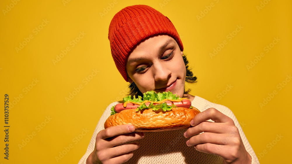 Young man in red beanie hat holding hot dog with lettuce and looking with pleasure, set against bright yellow background. Concept of street food, winter fairs, pop art, holiday snacks Young man in red beanie hat holding hot dog with lettuce and looking with pleasure, set against bright yellow background. Concept of street food, winter fairs, pop art, holiday snacks