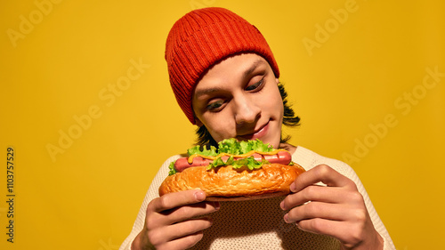 Young man in red beanie hat holding hot dog with lettuce and looking with pleasure, set against bright yellow background. Concept of street food, winter fairs, pop art, holiday snacks photo