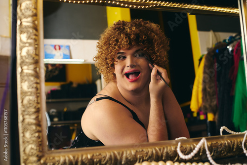 A drag queen applies makeup in a bustling backstage dressing room filled with holiday cheer.
