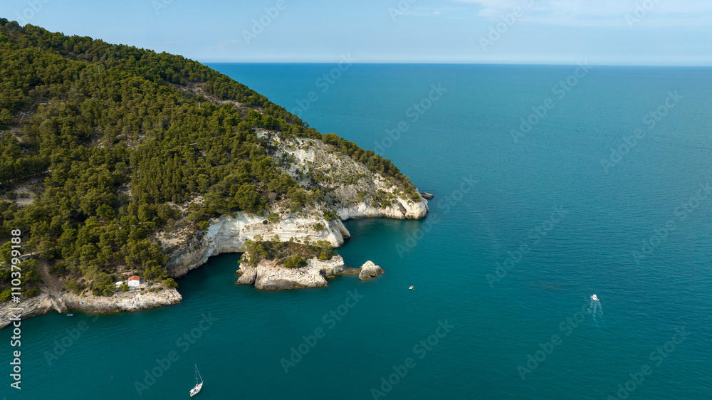 Aerial view of a hill on the Mediterranean Sea. There are boats on the surface of the crystal clear blue water.