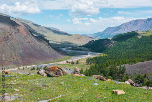 Vivid green landscape with shadows of clouds on wide alpine valley. Scenic sunny top view from grassy pass with big stones to long mountain river among coniferous trees. Lichen on boulder in sunlight.