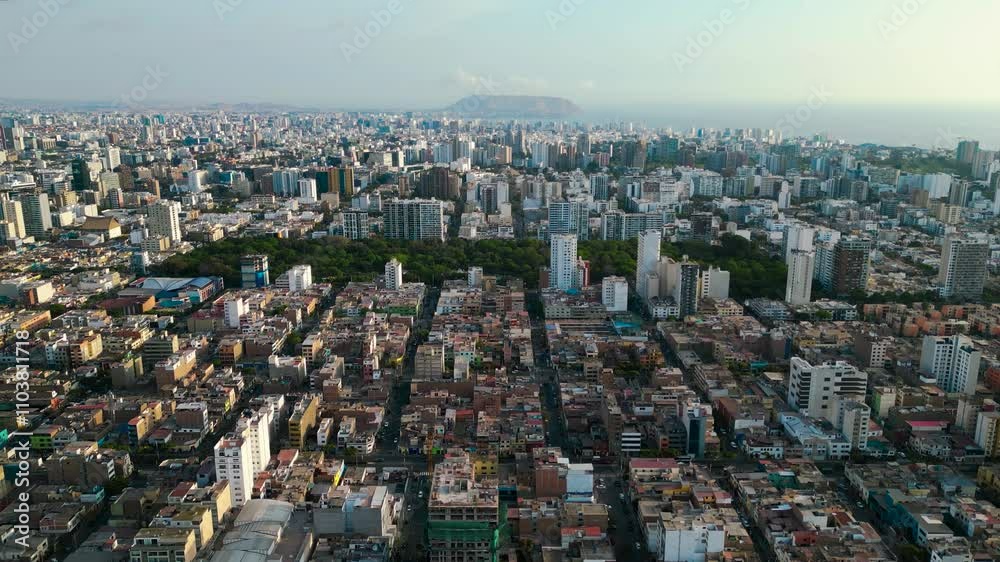 Toma panorámica con drone de la ciudad de Lima, con un exuberante parque verde y frondoso en el centro y el mar al fondo, donde se destaca un imponente peñasco, en una tarde de verano tranquilo