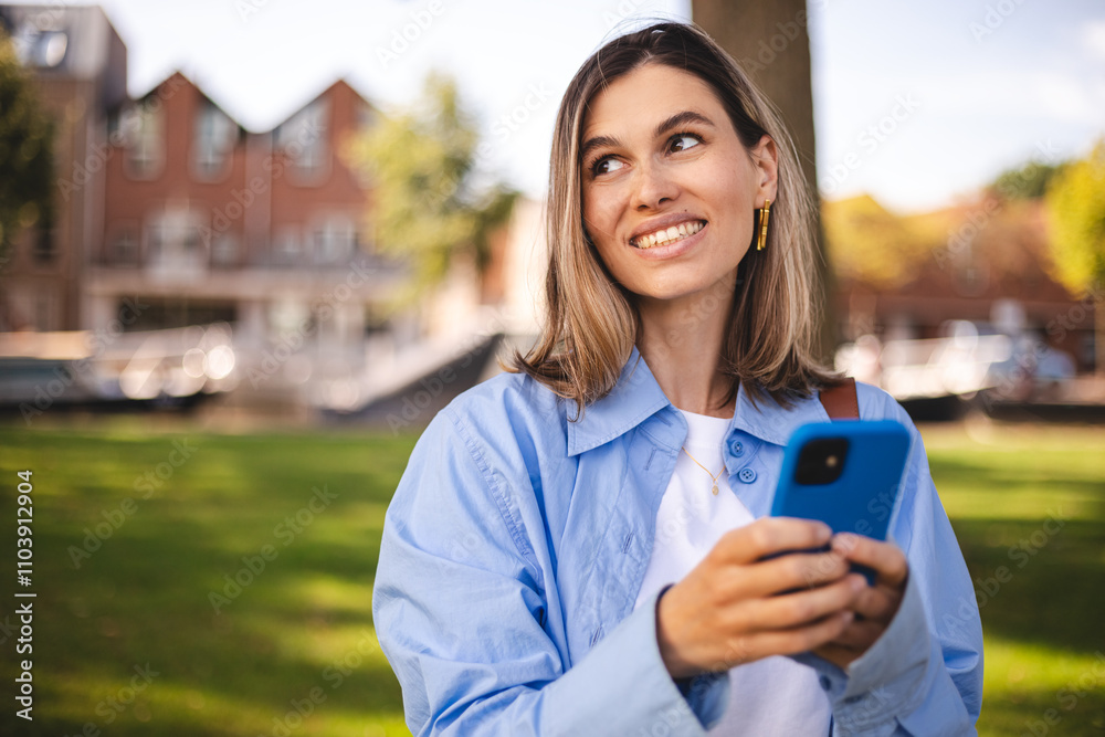 Attractive blonde short hair woman wear blue shirt hold mobile phone and look happy and smiling. Amazing enjoy woman walk in the park. Girl scrolling, messaging, write sms, read news, think answer. Attractive blonde short hair woman wear blue shirt hold mobile phone and look happy and smiling. Amazing enjoy woman walk in the park. Girl scrolling, messaging, write sms, read news, think answer.