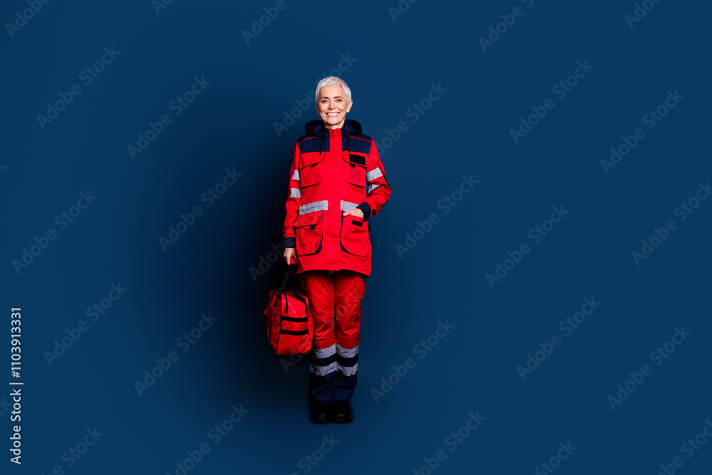 Full body photo of attractive senior woman ambulance worker first aid kit bag dressed red uniform isolated on dark blue color background Full body photo of attractive senior woman ambulance worker first aid kit bag dressed red uniform isolated on dark blue color background