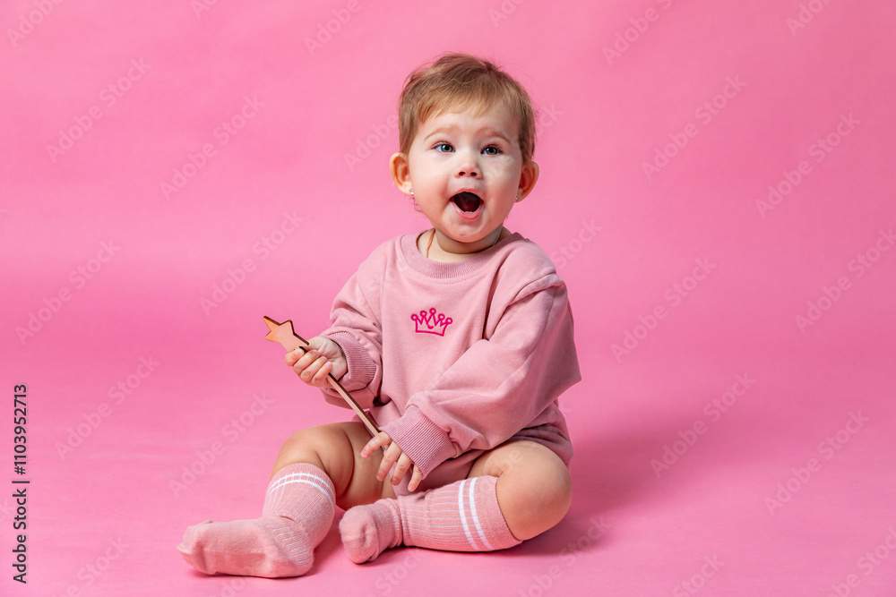 Surprised baby girl in a pink princess bodysuit sitting on a white background, holding magic wand. closeup. Childhood. First birthday party concept. High quality photo. Copy space Surprised baby girl in a pink princess bodysuit sitting on a white background, holding magic wand. closeup. Childhood. First birthday party concept. High quality photo. Copy space