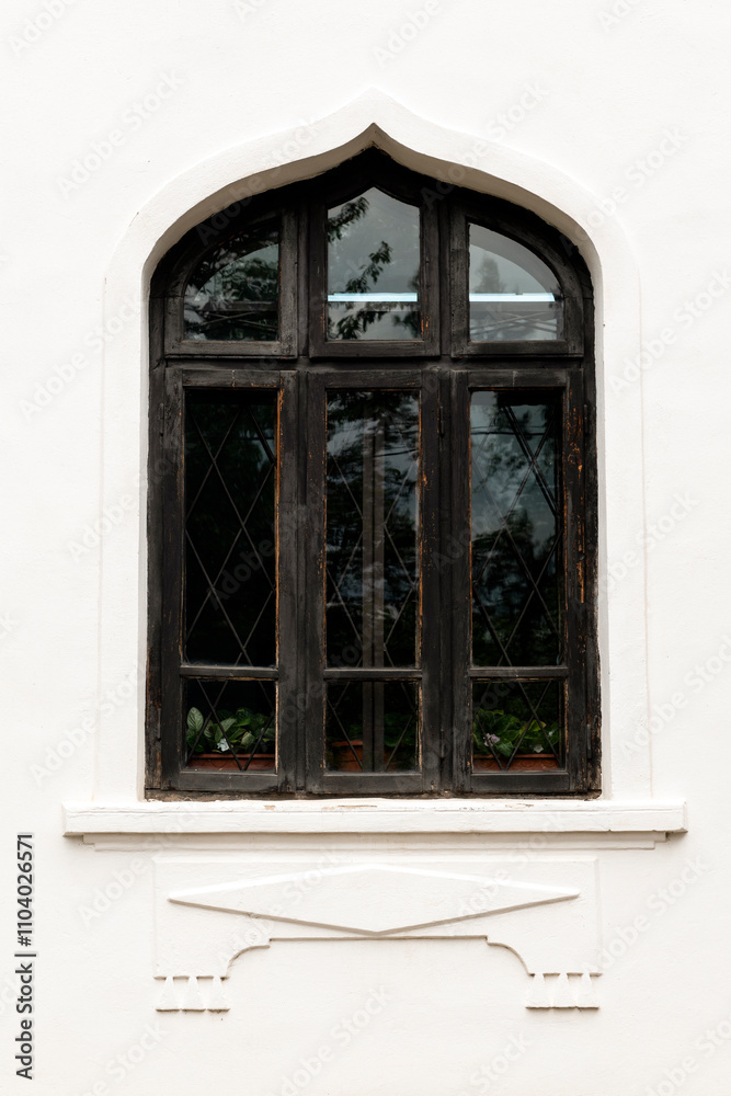 Fototapeta premium Vintage Arched Window Design. Elegant black arched window with intricate detailing on a white textured wall, reflecting surrounding greenery and sky, showcasing a timeless architectural style.