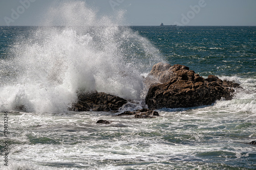 Powerful ocean waves collide with large rocks along the coast under a clear sky.