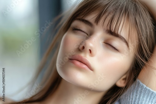 A serene young woman with long hair and soft features is resting with her eyes closed. She enjoys a moment of tranquility indoors, illuminated by gentle natural light, creating a peaceful atmosphere.