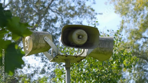 Outdoor warning siren speakers mounted on a tall pole, set against a bright blue sky and green foliage in 4K slow motion 120fps