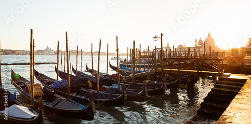 Panoramic view of a dock with gondolas tied to wooden posts and a silhouette of a church dome in the background with a romantic sunset in Venice, Italy