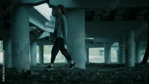 Full lowkey shot of young lady walking under bridge and suspicious masked man pursuing her and looking around, human trafficking