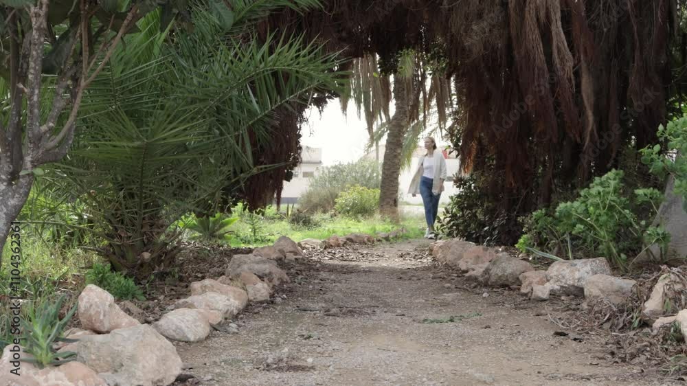Young woman is walking toward the camera on a path in a park. She is passing under the leaves of palm trees that are forming a natural archway Young woman is walking toward the camera on a path in a park. She is passing under the leaves of palm trees that are forming a natural archway