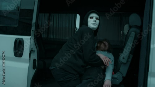 Tilt handheld shot of masked male criminal in black clothes carrying female victim and putting her inside van, looking around while committing crime, human abduction