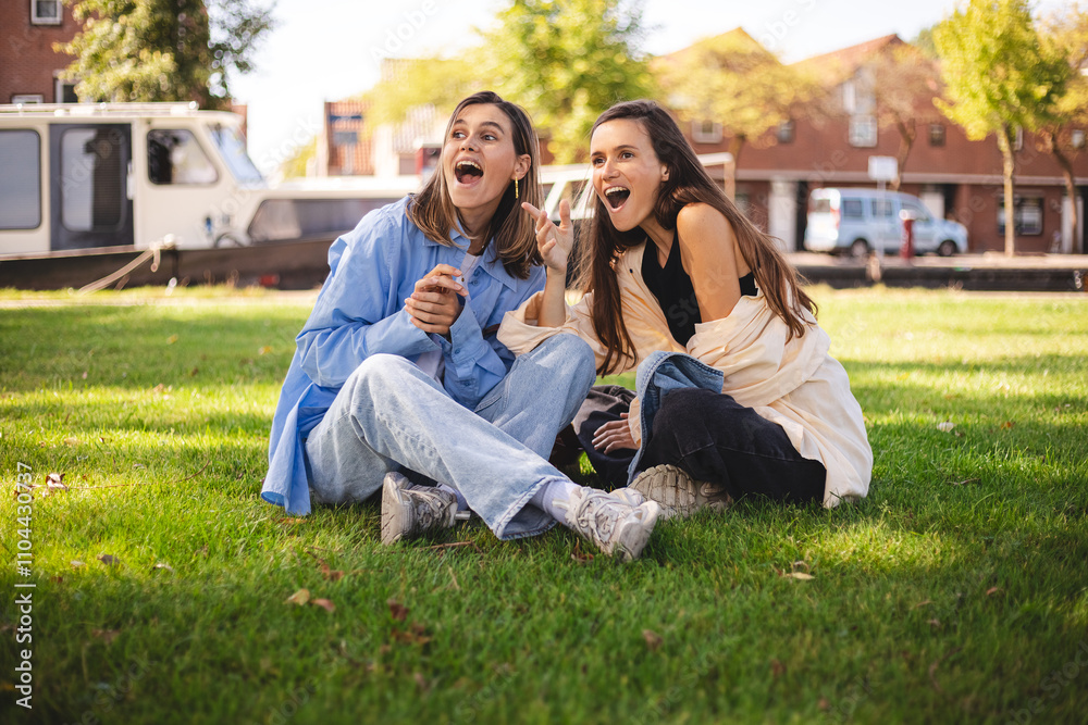 Two happy women friends sisters sitting in park on grass look at side point finger, laughing and show wow emotions. Two happy women friends sisters sitting in park on grass look at side point finger, laughing and show wow emotions.