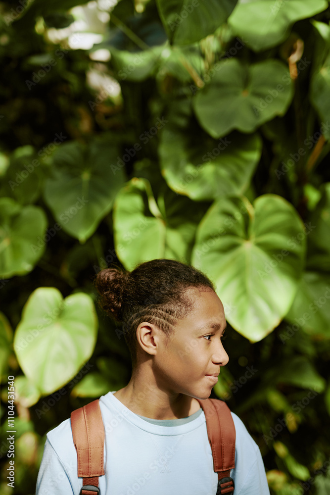 Vertical shot of African American teenager wearing backpack standing against green plant with big leaves in botanical garden Vertical shot of African American teenager wearing backpack standing against green plant with big leaves in botanical garden