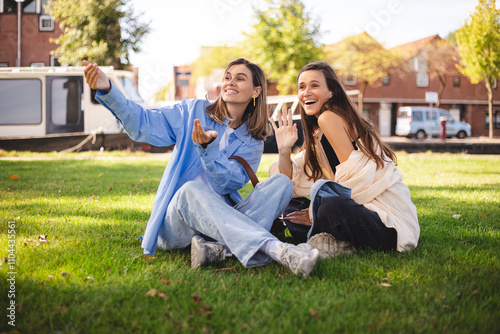 Fun stylish two women sisters friends waving a greeting with happy vivacious smile as they relax sitting together on grass in summer park. Happy girls meet they friends, welcome, come to us, hello, hi