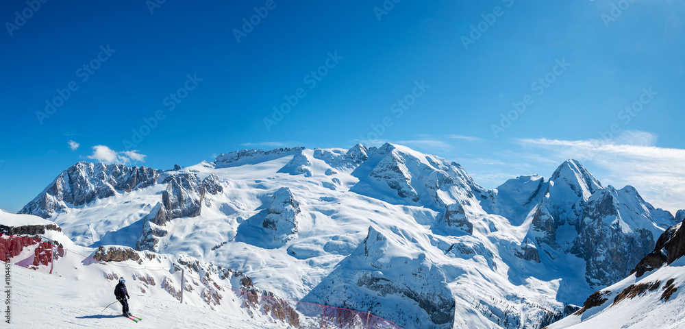 Marmolada mountain group and glacier in Dolomites in Italy in winter vied from Porta Vescovo in Arabba ski resort. Ski slope with red safety net and an unrecognizable skier. Blue sky with copy space
