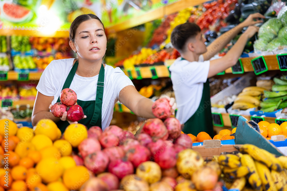 Happy young European woman in apron selling fresh pomegranate and fruits in hypermarket Happy young European woman in apron selling fresh pomegranate and fruits in hypermarket
