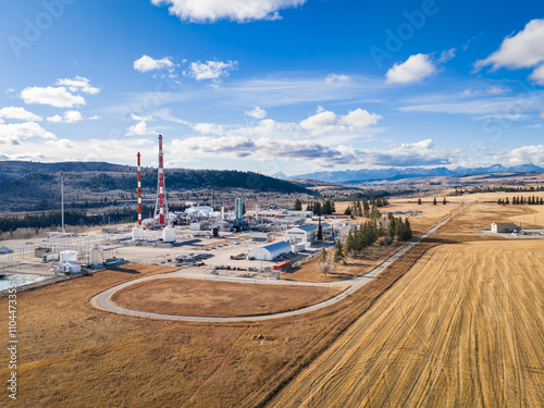 Aerial distant gas plant overlooking farm fields and hillside with Canadian Rocky Mountains at background near Cochrane Alberta Canada.
