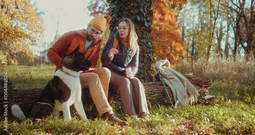 Happy hiking young couple with a dog taking a break sitting on a fallen tree in the forest Happy hiking young couple with a dog taking a break sitting on a fallen tree in the forest