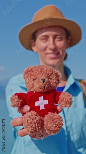 Vertical video. Woman in hat holding a small soft teddy bear with the image of the Swiss flag as a symbol of Switzerland against the background of the beautiful lake Interlaken in the city of Brienz photo