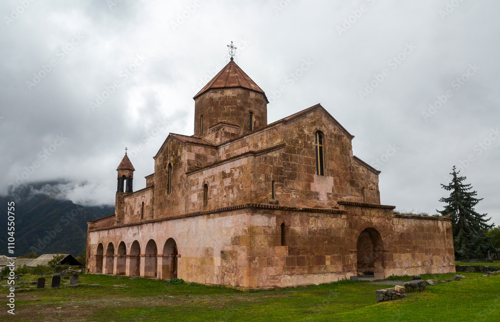 Historic Odzun church stands under a cloudy sky, surrounded by lush green grass and distant mountains, evoking a sense of serenity and timeless beautyю. Lori Region, Armenia