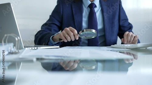 Businessman analyzing financial data with magnifying glass and calculator, conducting audit or review in office. Audit and taxes theme in business