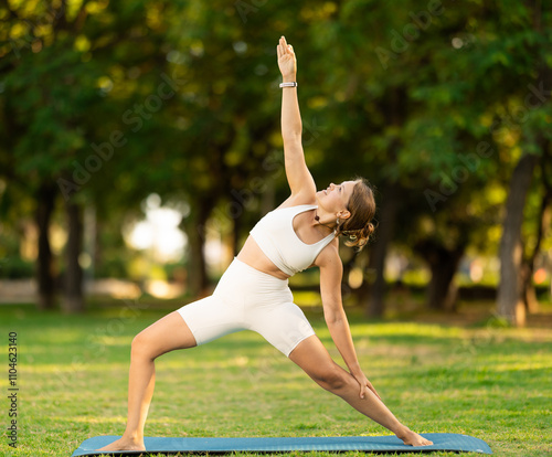 Sporty young girl performing standing lunging asana Viparita Virabhadrasana, or Reverse Warrior pose with backbend during yoga routine outdoors in green summer park photo