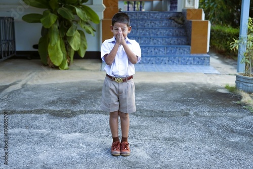 A Thai boy wearing a white school uniform raises his hands to say hello.