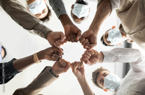 Success And Teamwork Concept. Below view of young diverse group of business people in protective medical masks making fist bump standing in circle. Workers do fist pump together celebrating good deal