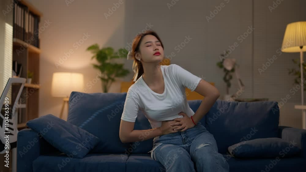 Young woman is experiencing intense stomach pain while sitting and then lying on her couch at night Young woman is experiencing intense stomach pain while sitting and then lying on her couch at night