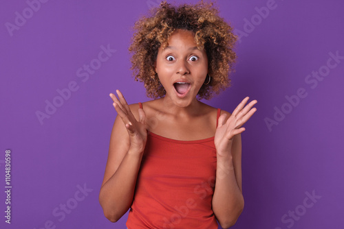 Papier peint Young African American female with curly hair shows excitement and surprise with wide eyes and raised hands against a vibrant purple background
