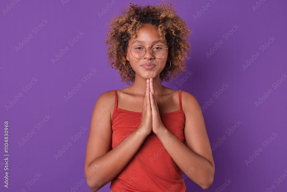 Joyful young African American woman stands in front of vibrant purple wall, her curly hair framing her face. She clasps her hands together in a gesture of appreciation, radiating positivity and warmth Joyful young African American woman stands in front of vibrant purple wall, her curly hair framing her face. She clasps her hands together in a gesture of appreciation, radiating positivity and warmth