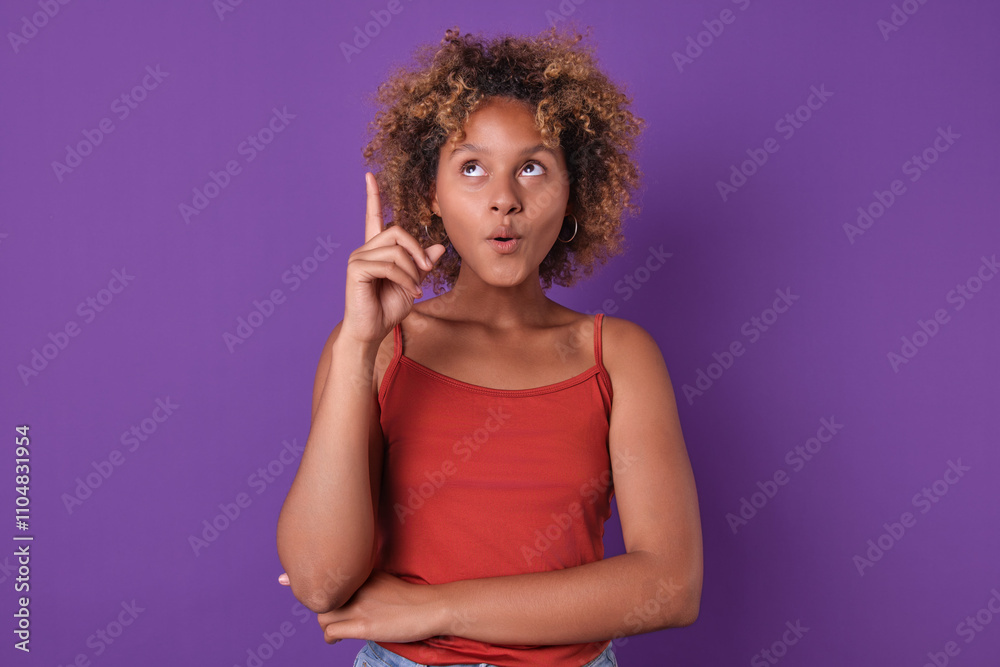 Young girl with curly hair is playfully pointing upwards with a curious expression. Dressed in a red top, she stands confidently against a bold purple wall, suggesting creativity and inspiration. Young girl with curly hair is playfully pointing upwards with a curious expression. Dressed in a red top, she stands confidently against a bold purple wall, suggesting creativity and inspiration.