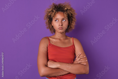 In striking purple setting, young African American woman with curly hair crosses her arms, wearing a red top. Her expression conveys mix of thoughtfulness and concern, creating an engaging atmosphere