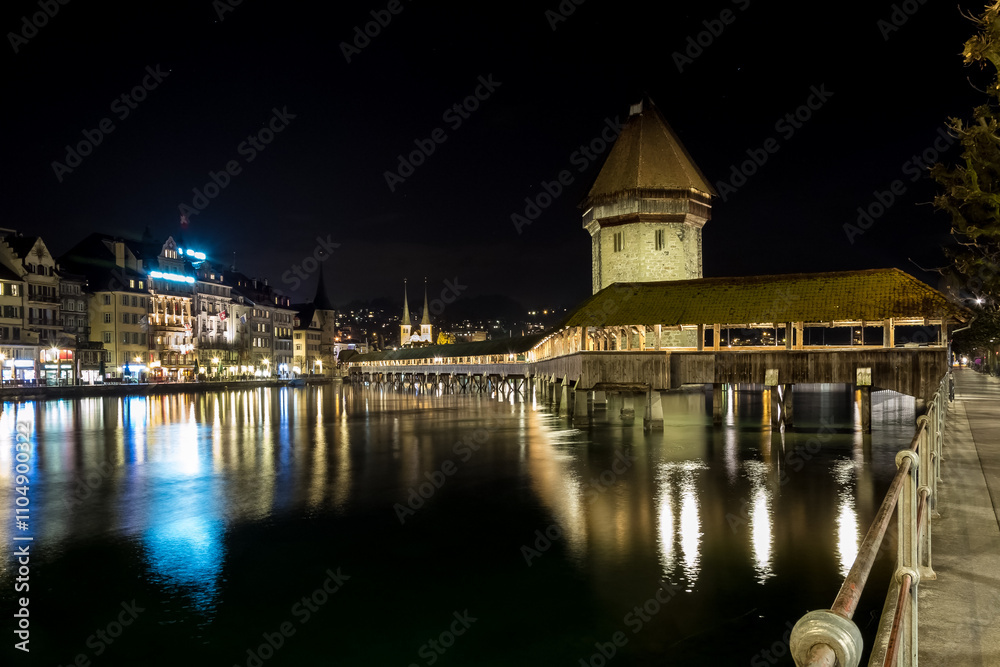 Fototapeta premium View of the Kapellbrücke (Chapel Bridge), a historic covered wooden footbridge and major landmark spanning the Reuss River, illuminated at night in the city of Lucerne, Switzerland.