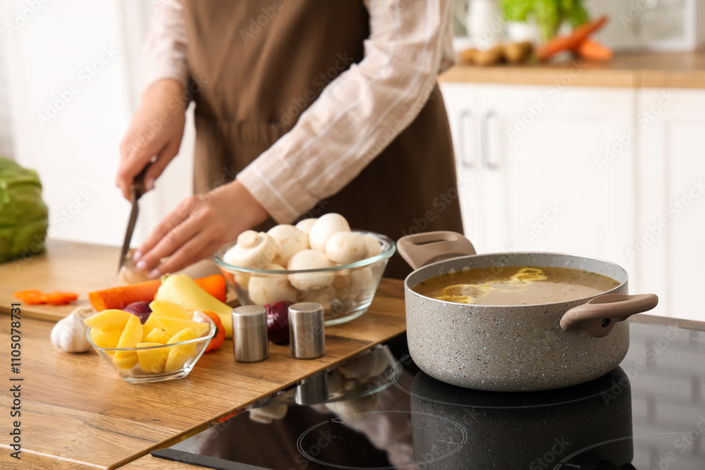 Cooking pot with soup on stove against woman cutting vegetables in kitchen, closeup Cooking pot with soup on stove against woman cutting vegetables in kitchen, closeup