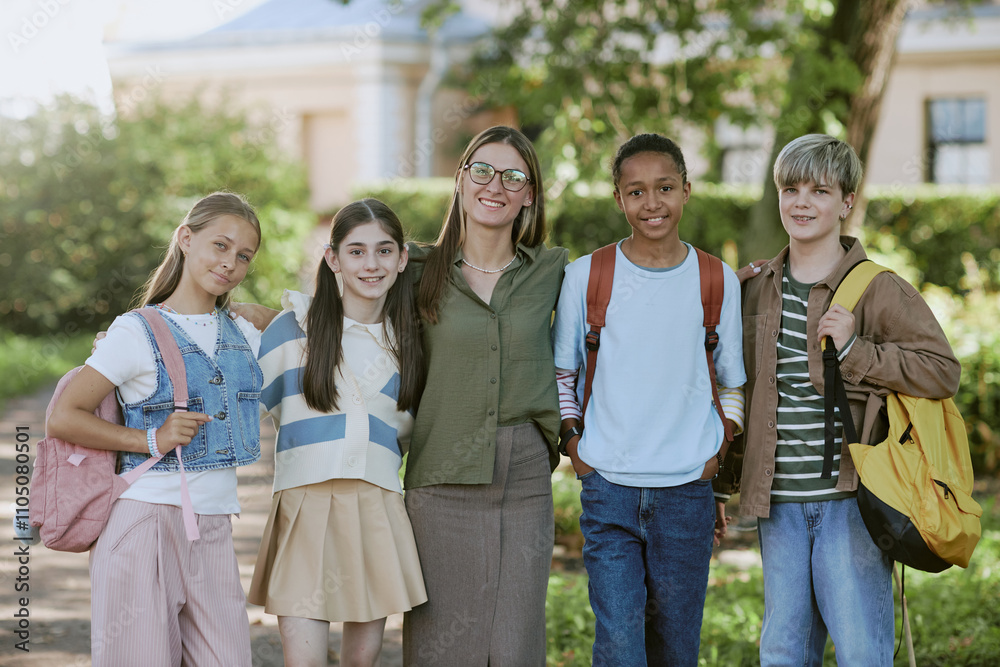 Group portrait of cheerful young female teacher and her teen students standing together in local park Group portrait of cheerful young female teacher and her teen students standing together in local park