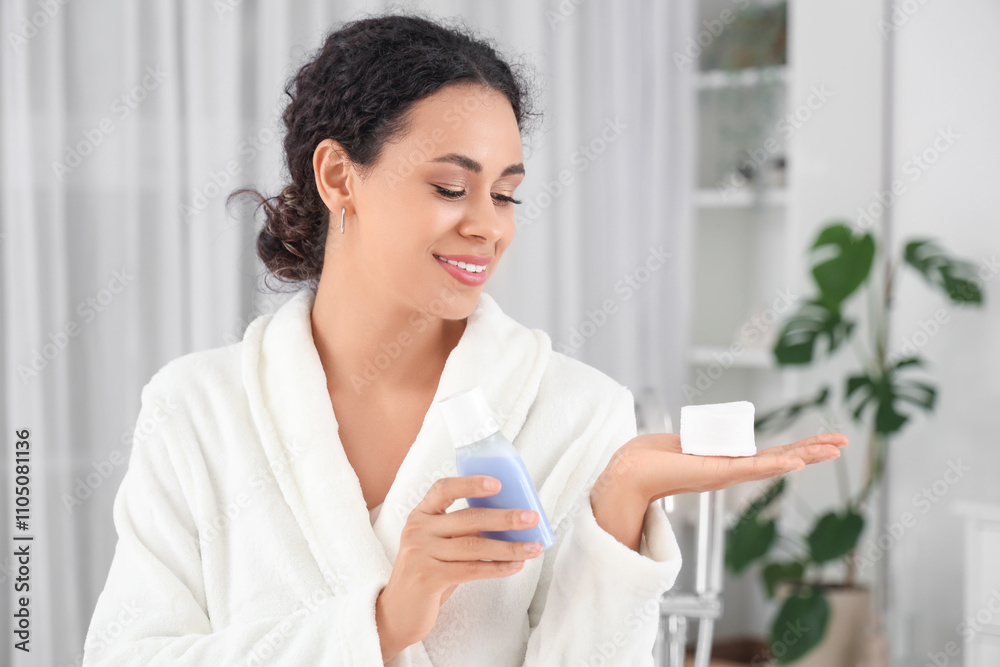 Beautiful young African-American woman with bottle of micellar water and cotton pads in bathroom Beautiful young African-American woman with bottle of micellar water and cotton pads in bathroom
