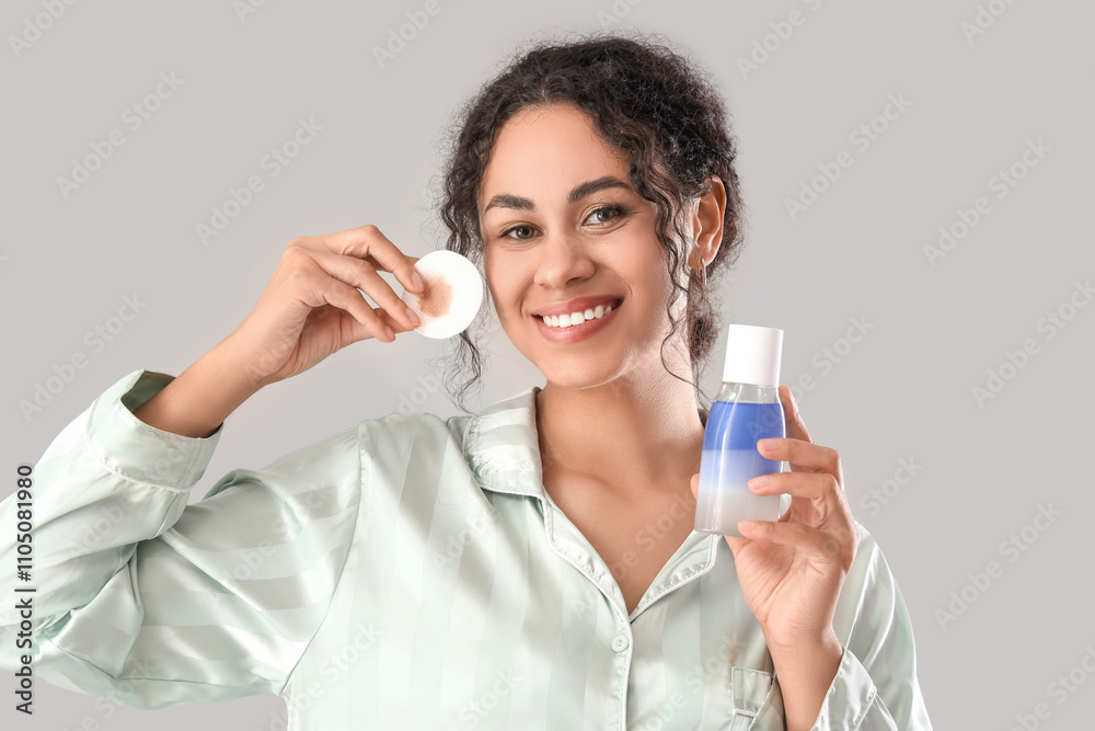 Happy young African-American woman with bottle of micellar water and cotton pad on grey background Happy young African-American woman with bottle of micellar water and cotton pad on grey background