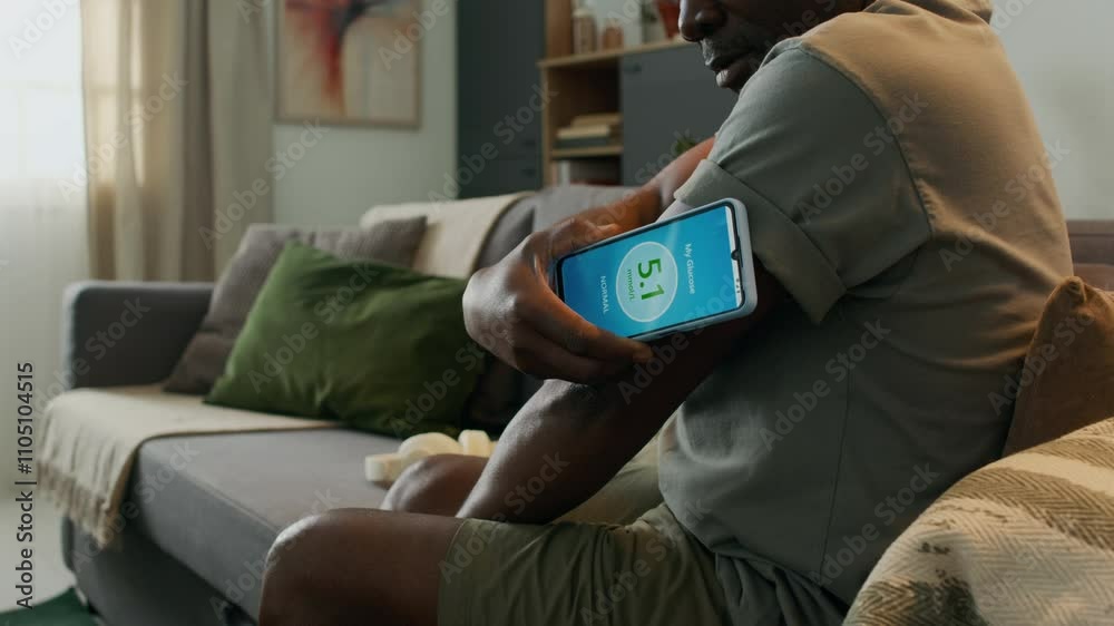 Close up view of Black man sitting on sofa in living room, scanning sensor of continuous glucose monitor with phone, then checking blood glucose level with mobile app