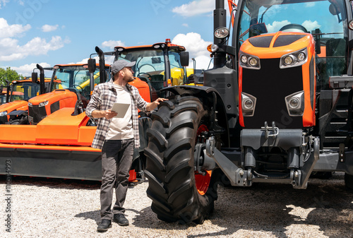 Caucasian man with digital tablet standing outdoors next to construction and agricultural machinery at dealership.