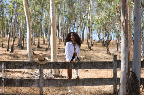 young, beautiful, Latin, brunette woman with curly hair wearing a white shirt, denim skirt and white boots rests one leg on the fence of the eucalyptus forest. The farmer is in the timber business. photo