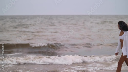 A woman in a white bikini and open shirt walks along a stormy beach under a gray sky. The dramatic weather and moody atmosphere evoke themes of solitude and introspection, perfect for conceptual and photo