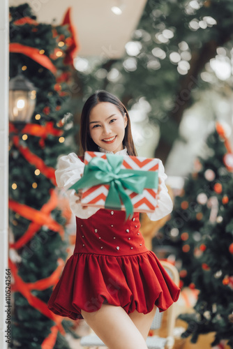 Christmas Cheer: A young woman beams with holiday joy, presenting a beautifully wrapped gift against a backdrop of twinkling Christmas trees.