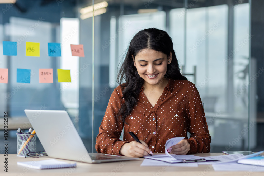 Hispanic woman businesswoman smiling while working at desk with laptop, papers, and pen. Engaging in productive work, showcasing focus and dedication to business tasks. Hispanic woman businesswoman smiling while working at desk with laptop, papers, and pen. Engaging in productive work, showcasing focus and dedication to business tasks.