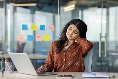 Hispanic woman businesswoman with neck pain sitting in office, laptop open, phone on table. Reflects modern work stress, need for ergonomic solutions, and digital life balance.