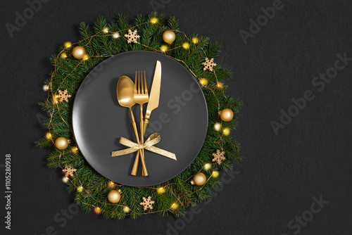 Black plate in a Christmas wreath with gold cutlery and decor on black background. Festive table setting. Copy space. Top view, flat lay.