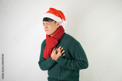 Young asian man wearing santa clause hat, red scarf and green long sleeved sweater  is suffering heart attack and holding his chest, isolated over white background.